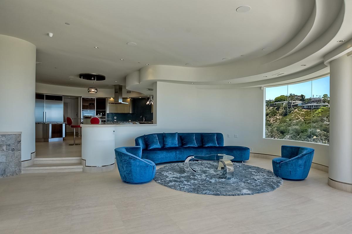 A light-filled living room with a minimalist interior inspired by cylindrical architecture. The space is not only large, but it is also open, with no doors separating it from other rooms. The contemporary sofa shape is a good match for the wall corner. In addition, the bold navy blue on velvet material stands out in this monochrome white space.