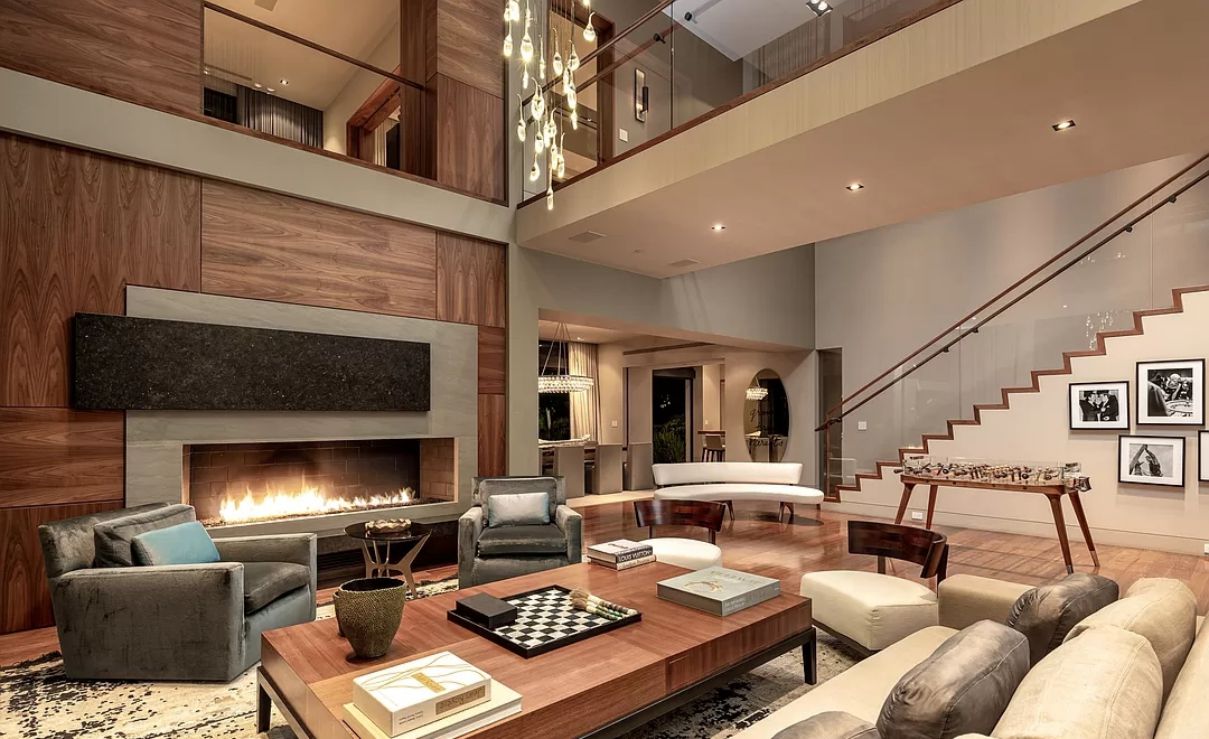 This contemporary living area is dominated by various colors of grey. Warm wood components like center tables, rafters, and the ceiling provide contrast to the interiors' black, white, and grey tones. The beautiful flower doddle wall feature and the distinctive metal pendant lighting are noteworthy features.