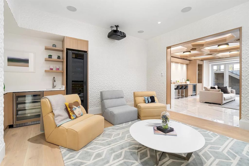 This is a light-filled living room with a white wooden ceiling with exposed wooden beams that matches the white walls and lets in an abundance of natural light via the surrounding large windows that illuminate the comfy black sofas.