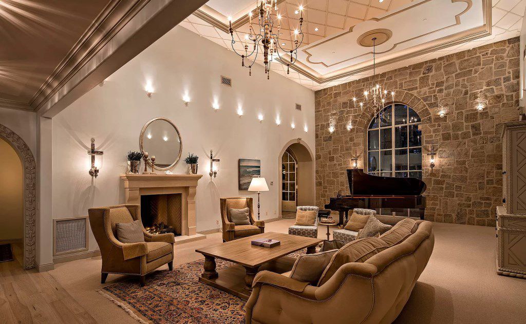 To the right of the room is a sizable stone fireplace with a variety of stone tile facings, and to the left are sizable stone French windows. The space is given the much-needed warmth it needs by the wooden high ceiling, gorgeous chandelier, and hardwood flooring. This living area is made cozier by the modern interpretation of the English sofa and the attractiveness of the wooden coffee tables. 