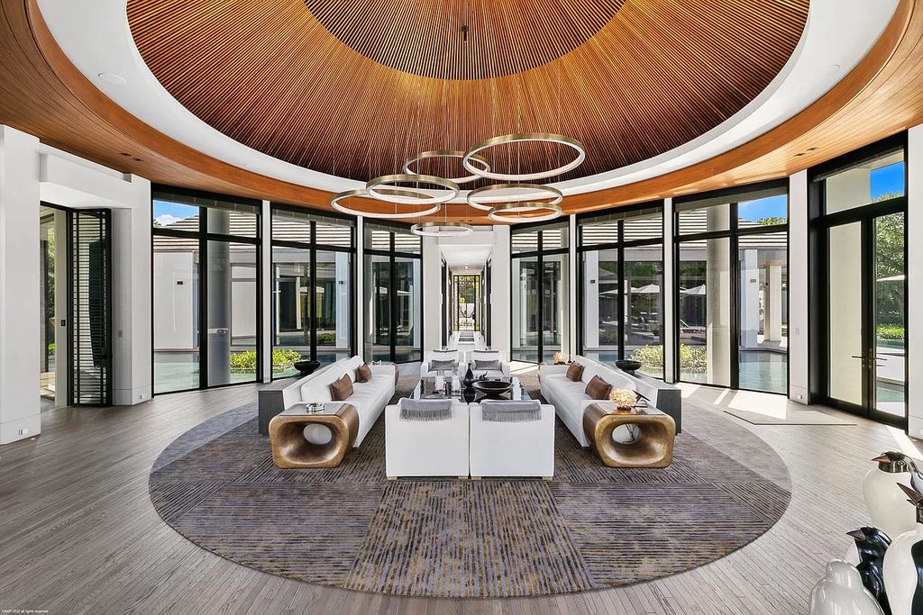 In the living room of the Mansion in Jupiter, the classic white sofa exudes relaxation and tranquility when placed at the center of the majestic and solemn space. The copper-plated metal wires of the ceiling stand out, perfectly synchronized with the chandelier design. The glass door system and the reflective metal furniture give the room a bright and airy atmosphere.